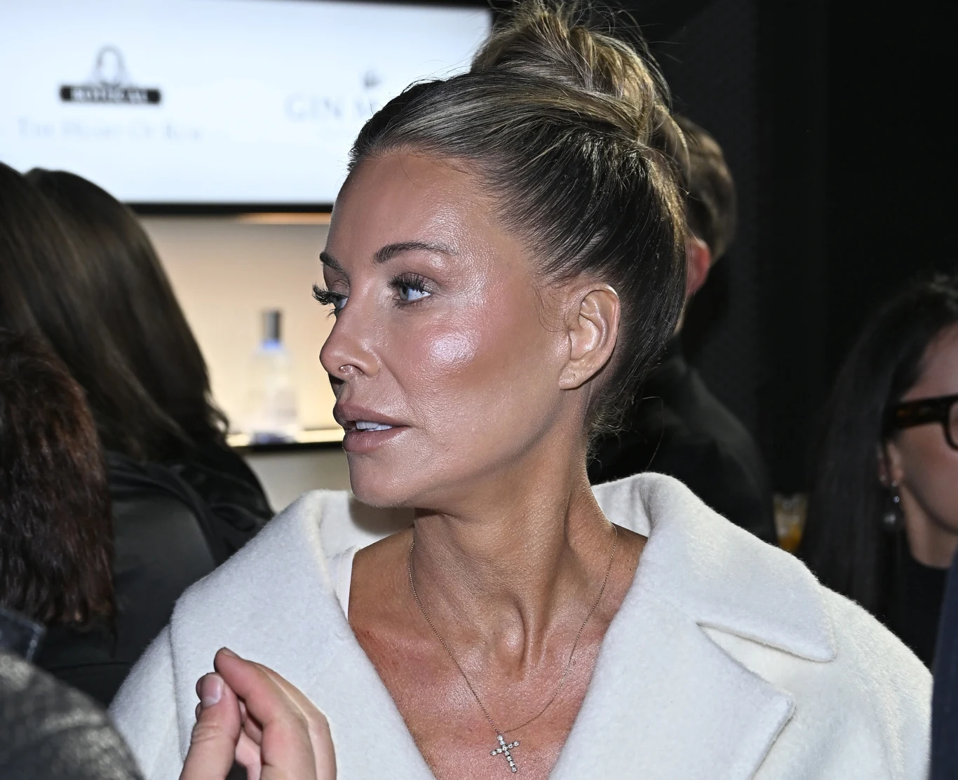 Kobieta o jasnych włosach upiętych w kok ubrana w jasny płaszcz, widoczna z profilu, na szyi delikatny naszyjnik z krzyżykiem, w tle osoby oraz rozmyty, jasny ekran.