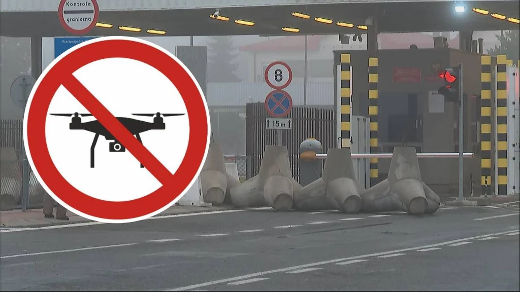 Zakaz używania dronów zaznaczony dużym znakiem graficznym na tle punktu kontrolnego zabezpieczonego betonowymi zaporami oraz szlabanem; oznaczenia drogowe i infrastruktura sugerują miejsce o podwyższonych środkach bezpieczeństwa.