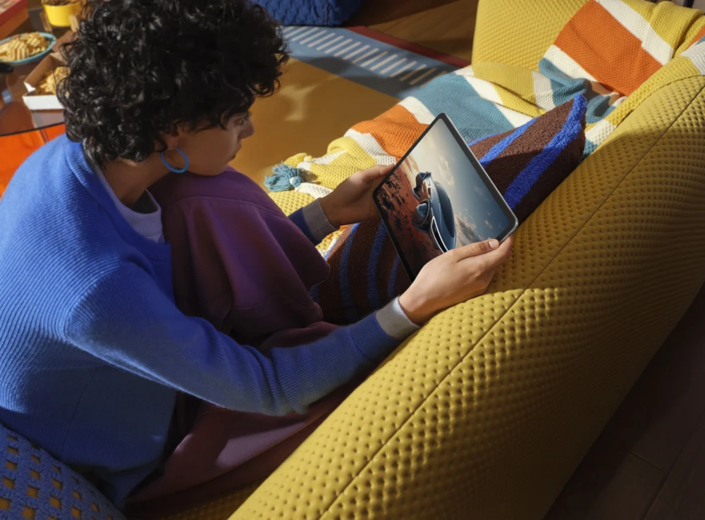 Osoba z kręconymi włosami siedzi na żółtej sofie, trzyma tablet i ogląda film lub zdjęcia, wokół niej kolorowy koc i poduszki, wnętrze jest jasne i przytulne.
