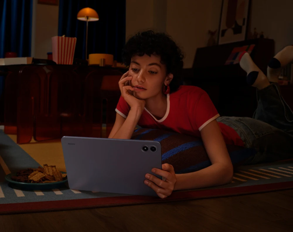 Młoda osoba leży na dywanie w pokoju, opierając głowę na dłoni i trzymając tablet. W tle widoczne miękkie światło lampy, przekąski i nastrojowy wystrój wnętrza.