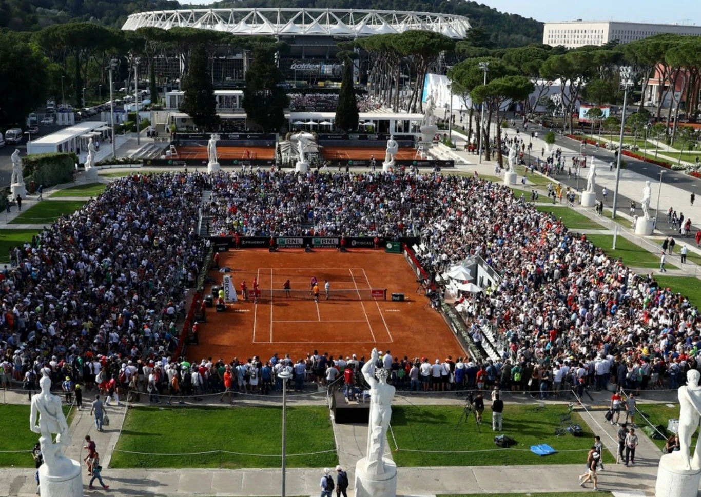 Stadion imienia Nicoli Pietrangeliego w Rzymie Kort tenisowy otoczony trybunami pełnymi kibiców, na którym toczy się mecz na ceglanej nawierzchni, w tle widoczny stadion oraz zabytkowe rzeźby i zieleń.