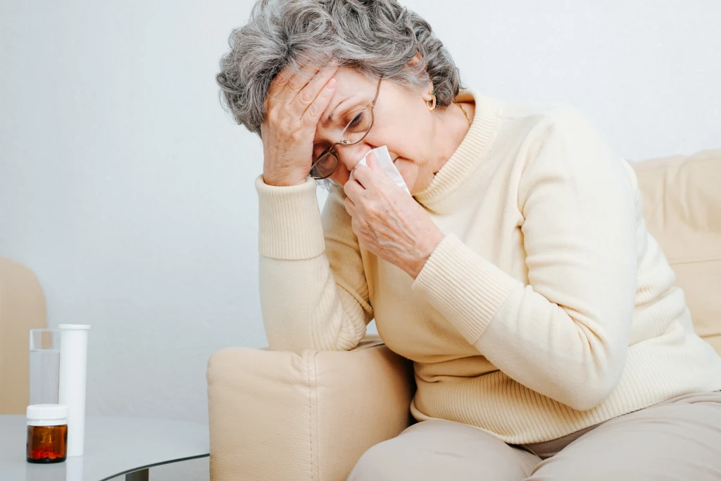 Częsty błąd może przedłużać infekcję i zwiększać ryzyko zakażenia bakteryjnego Starsza kobieta siedzi na beżowej sofie, trzyma chusteczkę przy nosie i podpiera głowę ręką, wyrażając oznaki złego samopoczucia lub przeziębienia. Na stoliku obok znajdują się lekarstwa i szklanka wody.