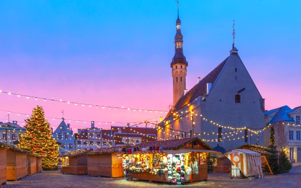 Świąteczny jarmark z drewnianymi straganami oraz dużą, oświetloną choinką na tle zabytkowego budynku z wysoką wieżą przy zachodzie słońca. Dopełnieniem sceny są liczne dekoracyjne światełka zawieszone nad placem.