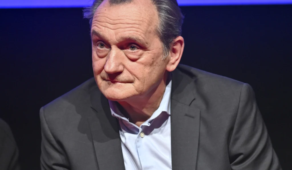 Starszy mężczyzna w ciemnym garniturze i jasnej koszuli siedzi na tle ciemnego, jednolitego tła z niebieskim pasem u góry kadru; ma poważny, zamyślony wyraz twarzy.