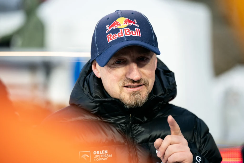 Adam Małysz Mężczyzna w czapce z logo Red Bull i kurtce sportowej spoglądający w stronę obiektywu, w tle nieostre elementy otoczenia.