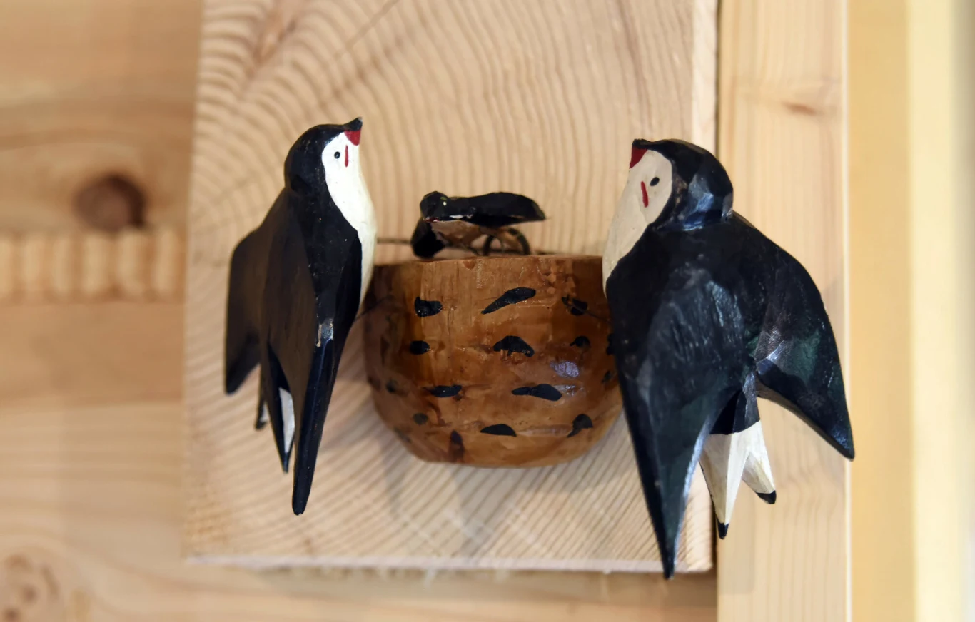 Dwie drewniane figurki jaskółek siedzące przy gnieździe wykonanym z drewna, ustawione na tle jasnej drewnianej ściany.