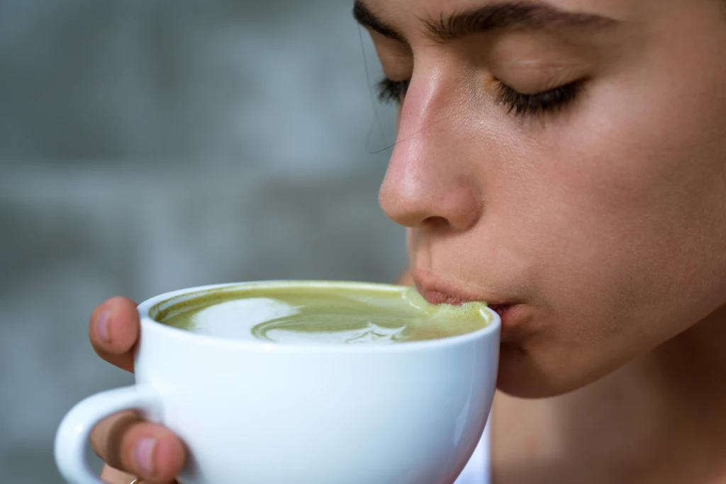 Jakie są alternatywy dla picia kawy? Młoda osoba trzyma białą filiżankę z zielonym napojem i delikatnie pije kawę matcha, mając zamknięte oczy i skupiony wyraz twarzy.