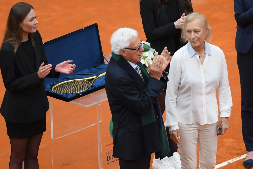 Ceremonia wręczenia nagrody na korcie tenisowym, kilka osób w eleganckich strojach, złota rakieta tenisowa umieszczona w otwartym etui na przezroczystym stole, kobieta w białej koszuli i mężczyzna w okularach klaskający.