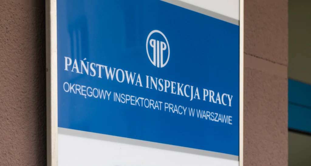 Tablica informacyjna Państwowej Inspekcji Pracy, Okręgowy Inspektorat Pracy w Warszawie, na tle fragmentu ściany budynku.