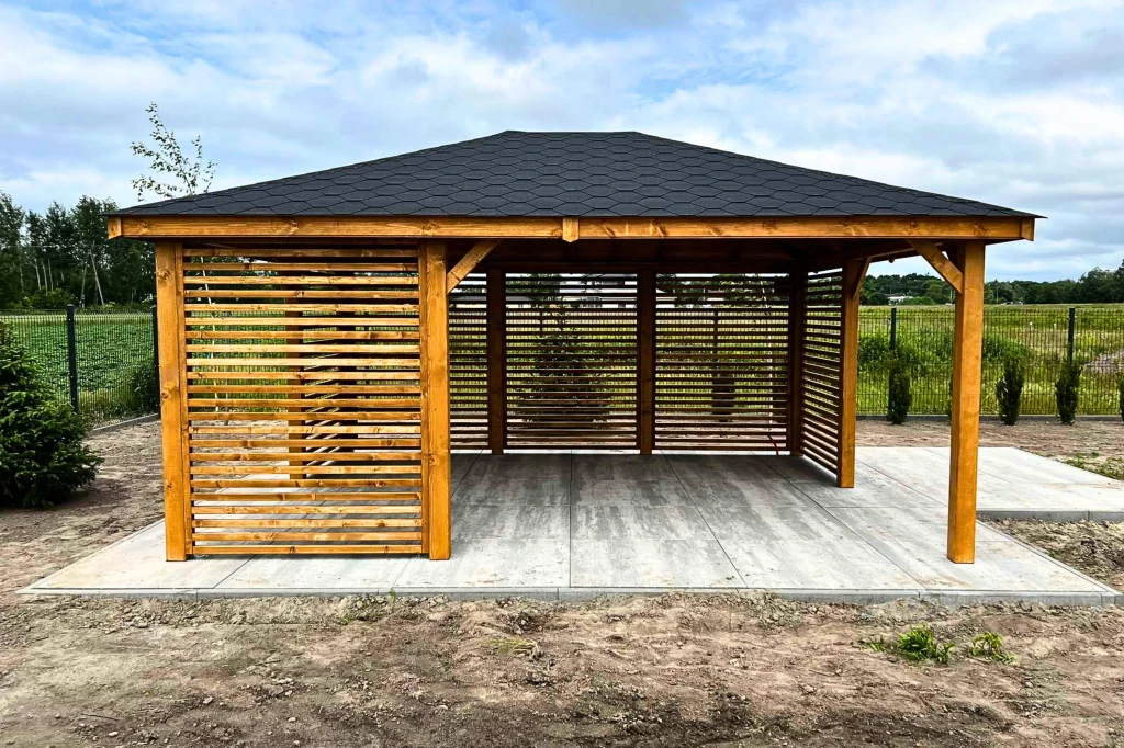 Drewniana altana ogrodowa z ażurowymi ścianami na betonowym podłożu, otoczona ogrodem z zielonym trawnikiem, żywopłotem i polami w tle.