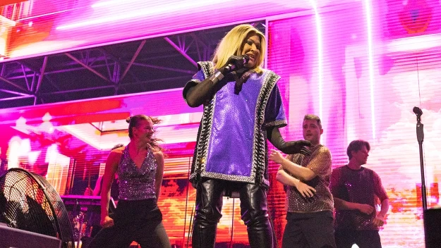 Wokalista w jasnej peruce i stroju sceniczno-błyszczącym stoi na scenie w otoczeniu tancerzy wykonujących dynamiczny układ choreograficzny, tło podświetlone kolorowymi światłami LED.