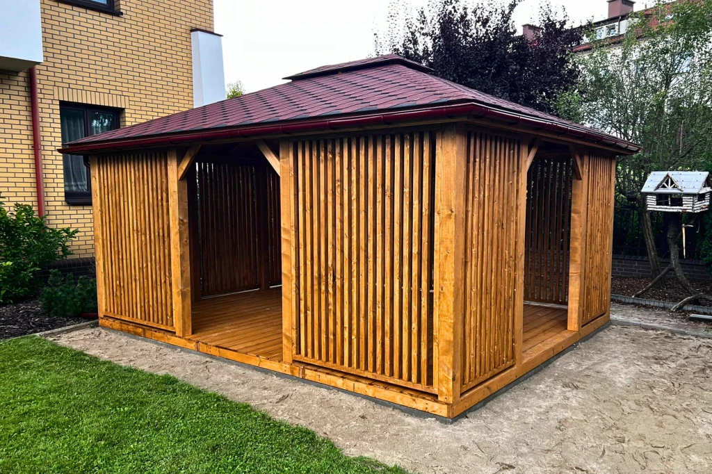 Drewniana altana ogrodowa z ażurowymi ścianami i dwuspadowym, czerwonym dachem, stojąca na podłożu gruntowym tuż przy trawniku, w pobliżu budynku mieszkalnego i zieleni.