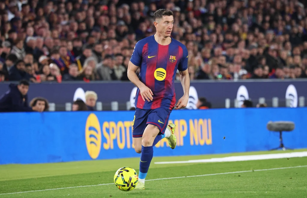 Robert Lewandowski w barwach FC Barcelona podczas sezonu 2025/2026 Piłkarz w stroju FC Barcelony biegnie z piłką po boisku podczas meczu, w tle widownia zapełniona kibicami oraz bandy reklamowe.