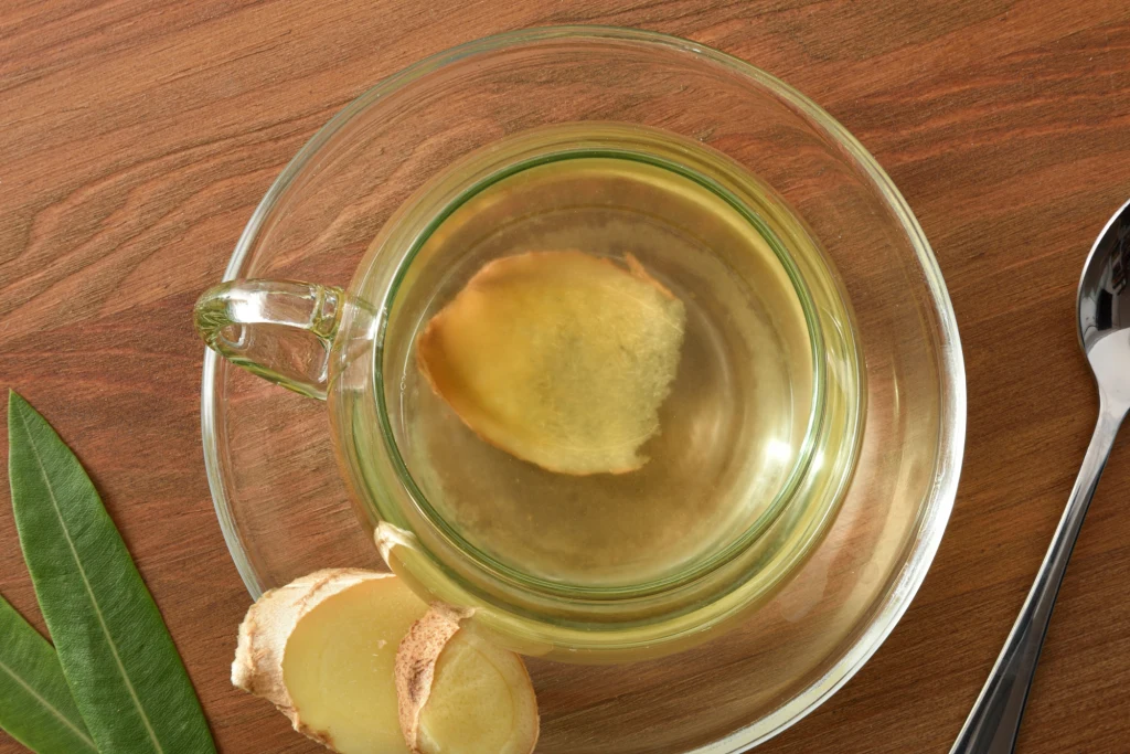 Zastąp tym napojem kawę Szklanka naparu z imbirem ustawiona na drewnianym blacie, obok plasterki świeżego imbiru i liść dekoracyjny, całość dopełniona metalową łyżeczką po prawej stronie.