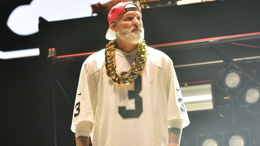 Brodaty mężczyzna w białej sportowej koszulce z dużą cyfrą 3 na piersi, z ogromnym złotym łańcuchem na szyi i czerwoną czapką z daszkiem założoną tył naprzód, stoi na scenie w świetle reflektorów.