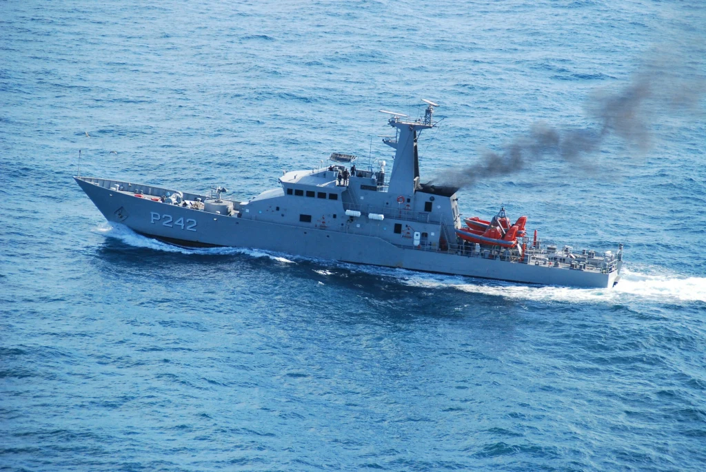 Wojskowy okręt patrolowy oznaczony numerem P242 płynie po spokojnej, niebieskiej wodzie, z komina wydobywa się czarny dym, na pokładzie widoczne są pomarańczowe łodzie ratunkowe.