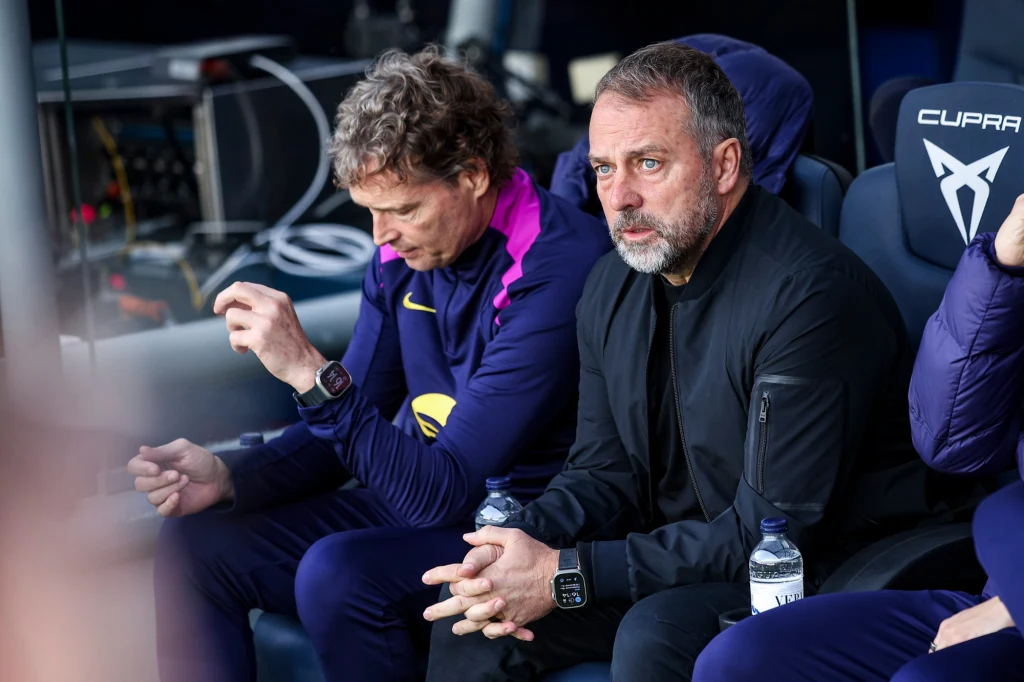 Hansi Flick poruszony po meczu z Alaves Dwóch mężczyzn siedzących na ławce trenerskiej podczas wydarzenia sportowego, skupionych i zamyślonych, ubranych w sportowe stroje. Przed nimi butelki wody, w tle widać fragment logo CUPRA.