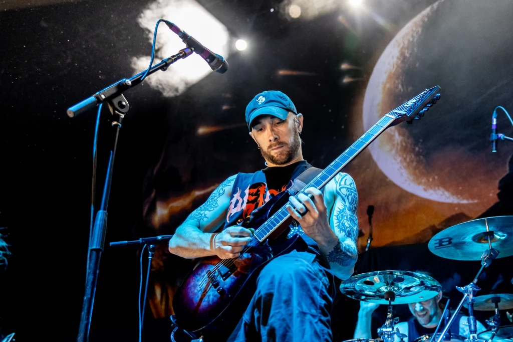 Josh Middleton (Sylosis) w akcji Gitarzysta podczas koncertu, intensywnie grający na gitarze elektrycznej, z widocznymi tatuażami na rękach i wyrazistą mimiką twarzy, w tle perkusista oraz scenografia z motywem kosmicznym.
