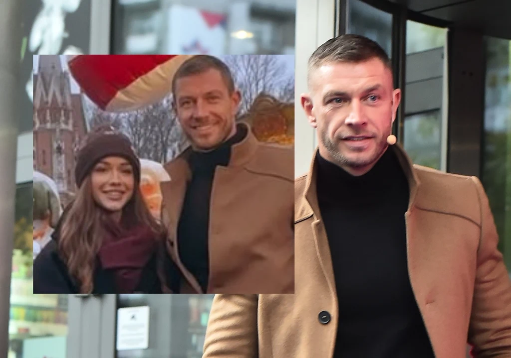 Mężczyzna ubrany w beżowy płaszcz oraz czarny golf stoi przed wejściem do nowoczesnego budynku, na drugim planie widać jego zdjęcie z uśmiechniętą kobietą, oboje ubrani elegancko, w tle fragment zabudowy miejskiej i duża czerwona dekoracja.