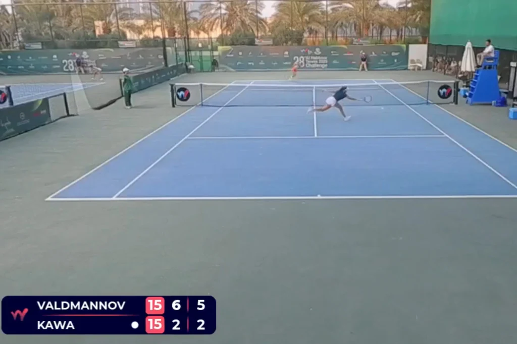 Tak Vendula Valdmannova mijała Katarzynę Kawę Kort tenisowy z zawodniczką wykonującą dynamiczny ruch do piłki, otoczenie boiska z widoczną zielenią oraz tablicą wyników w lewym dolnym rogu ekranu.