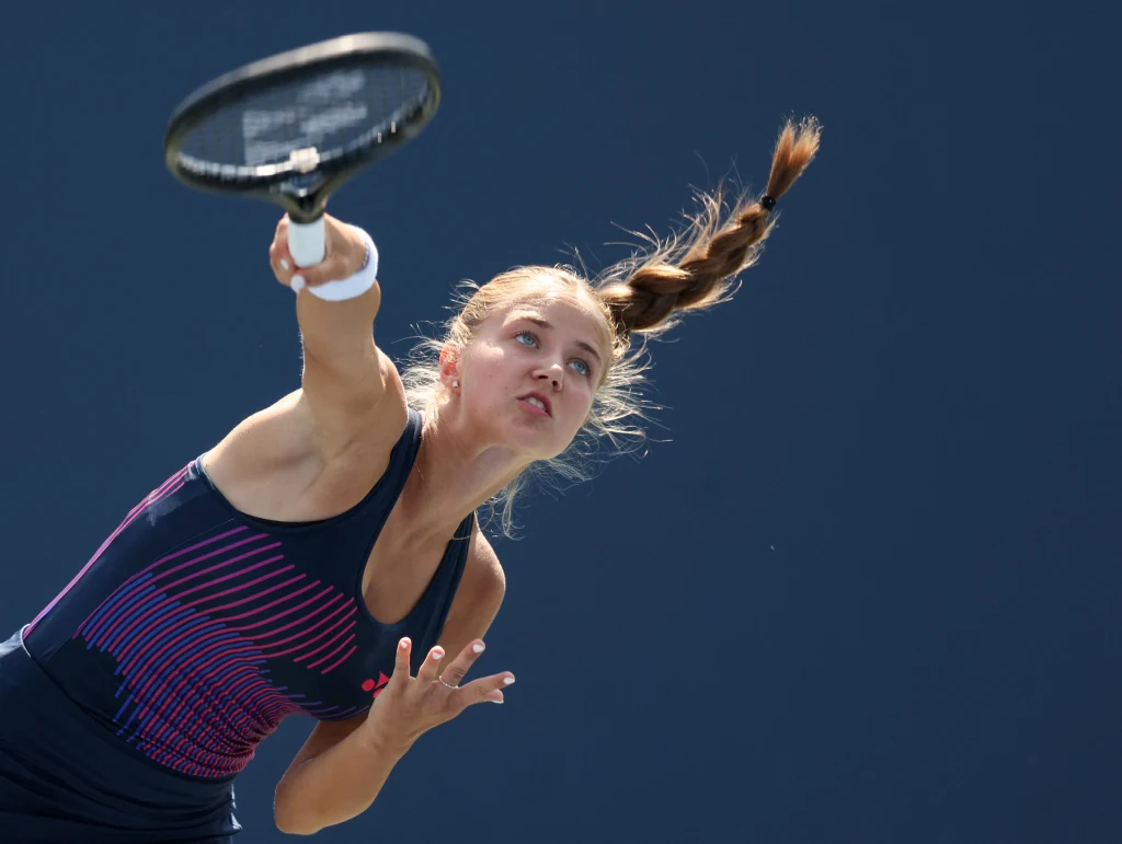 Vendula Valdmannova Młoda kobieta w sportowym stroju wykonuje serwis tenisowy, dynamiczny ruch i skoncentrowane spojrzenie podkreślają energię oraz zaangażowanie zawodniczki, tło jest jednolite i nieodwracające uwagi od głównego tematu.