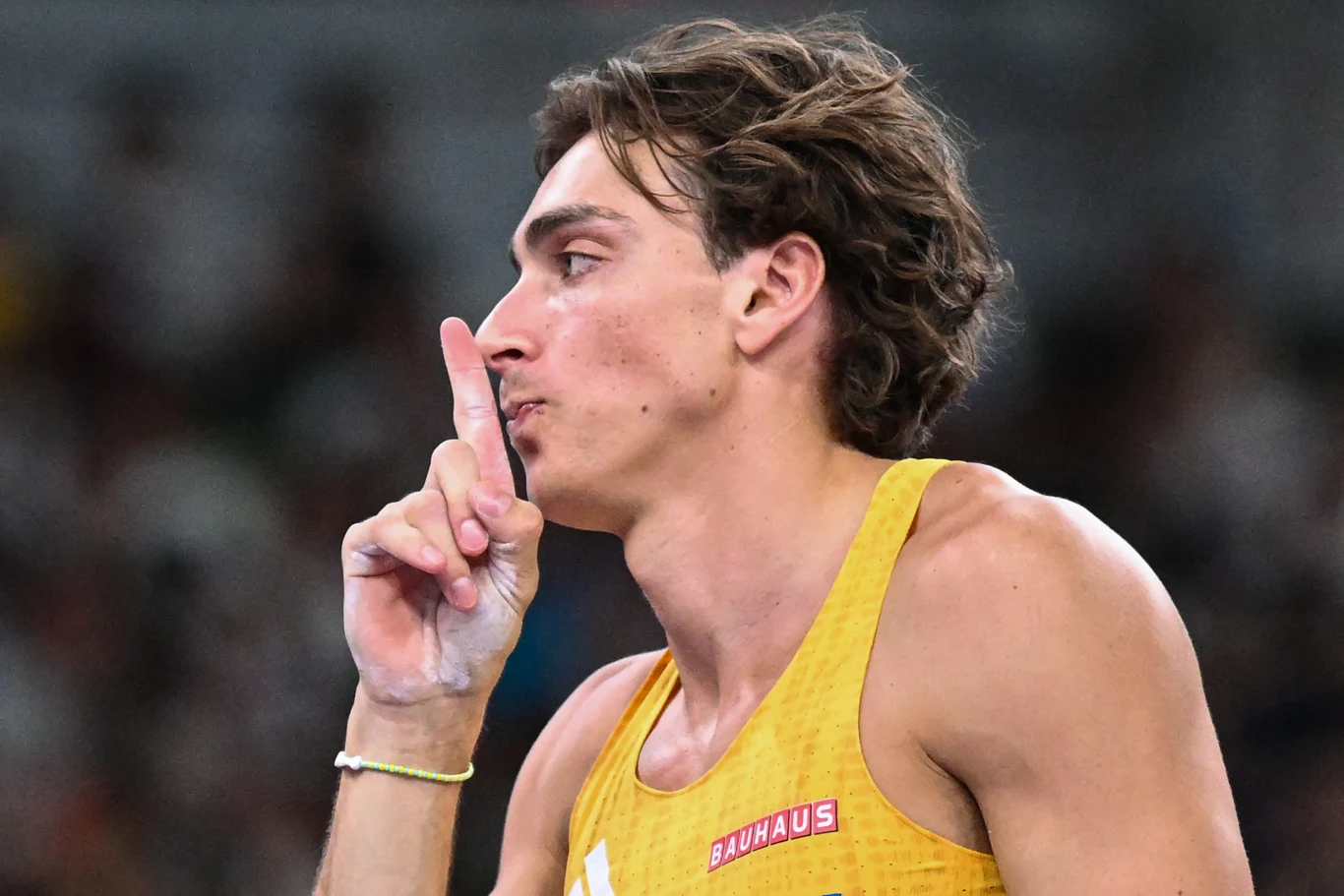 Mężczyzna w żółtym stroju sportowym z brązowymi włosami trzyma palec wskazujący przy ustach, wykonując gest ciszy. W tle rozmyte otoczenie stadionowe.