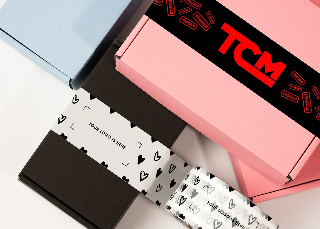 Kolorowe pudełka kartonowe ozdobione ozdobnymi taśmami z nadrukami, jedno z widocznym czerwonym logo TCM, inne z serduszkami i miejscem na personalizowane logo.