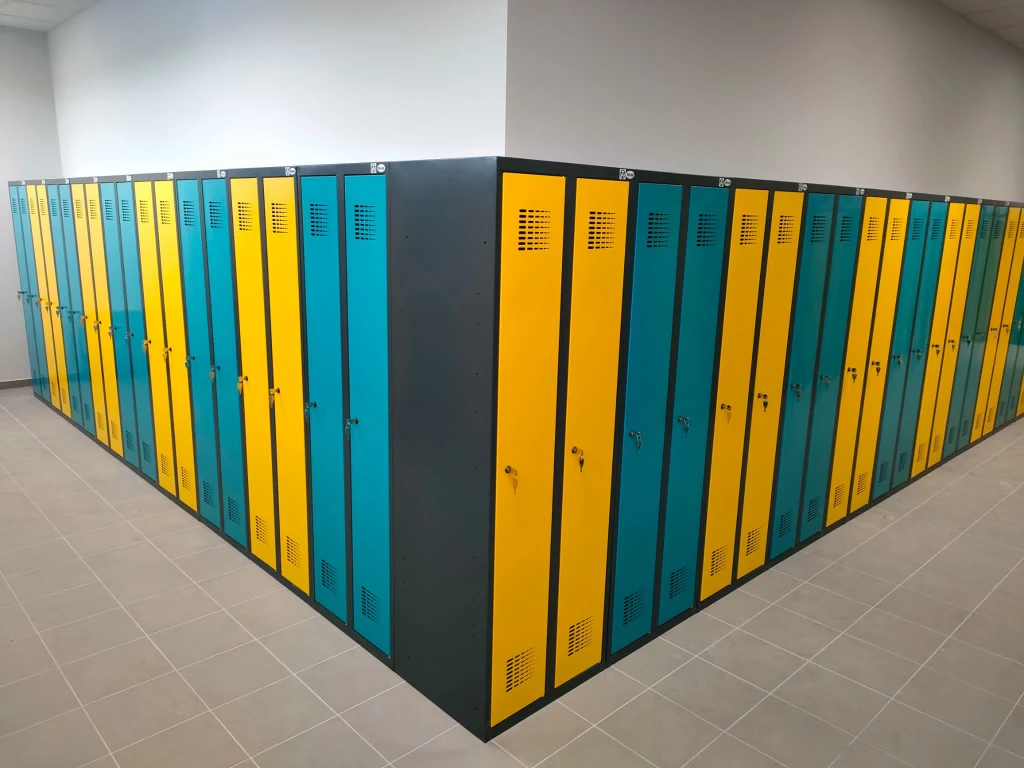 Rząd szafek metalowych w dwóch kolorach: niebieskim i żółtym, ustawionych pod kątem prostym w narożniku jasnego, wyłożonego kafelkami pomieszczenia.