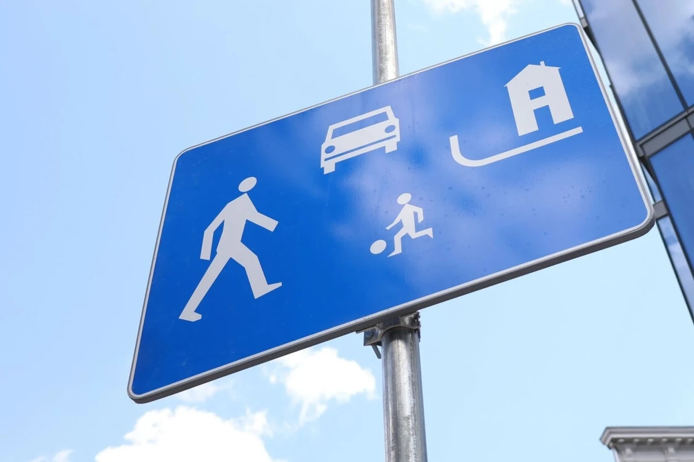 Wbrew obiegowym opiniom w strefie zamieszkania nie wolno parkować "gdzie się chce", a jedynie na wyznaczonych miejscach. Jakie jeszcze w niej panują zasady? Niebieska tablica drogowa z białymi piktogramami przedstawiającymi osobę dorosłą, dziecko z piłką, samochód oraz dom wskazująca strefę zamieszkania i pieszych.