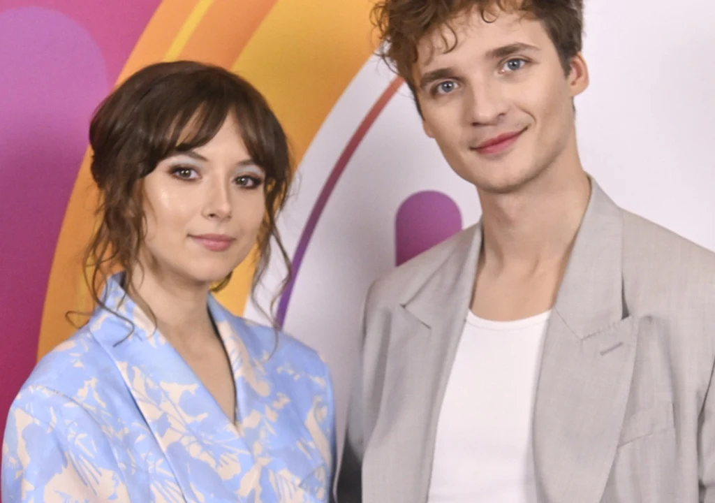 Kasia i Jacek Sienkiewiczowie Dwie młode osoby stoją obok siebie na tle kolorowej ściany z motywami geometrycznymi, ubrani w eleganckie stroje, kobieta w jasnoniebieskim garniturze, mężczyzna w jasnoszarej marynarce i białej koszulce, oboje patrzą z lekkim uśmiechem w kierunku apar...