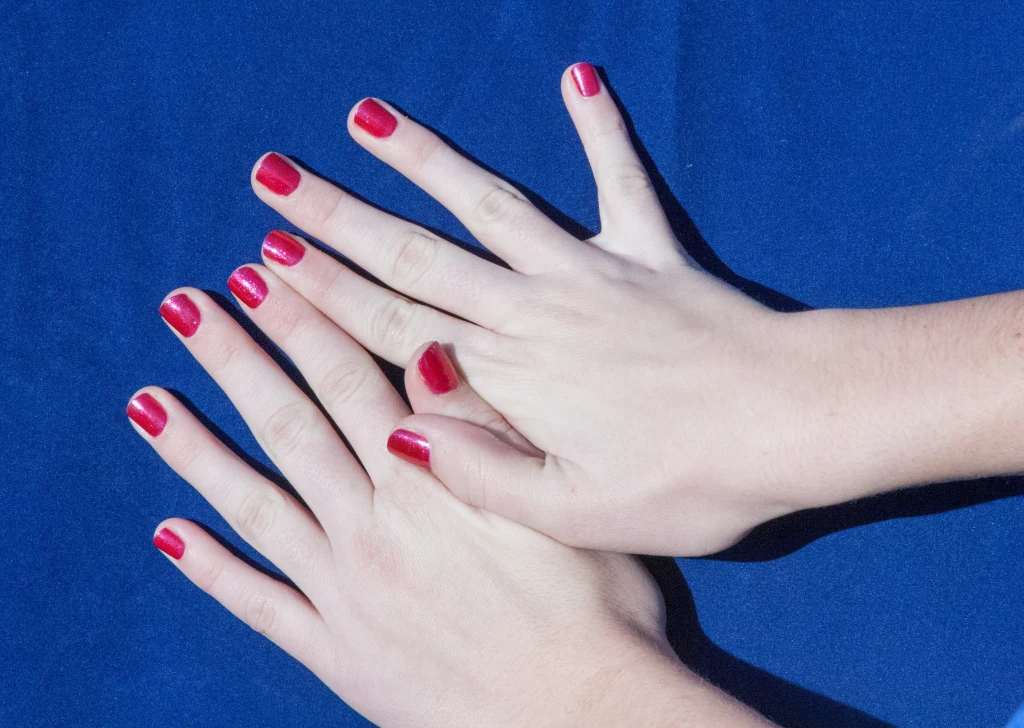 Dwie kobiece dłonie z zadbanymi paznokciami pomalowanymi na czerwono na tle granatowego materiału.