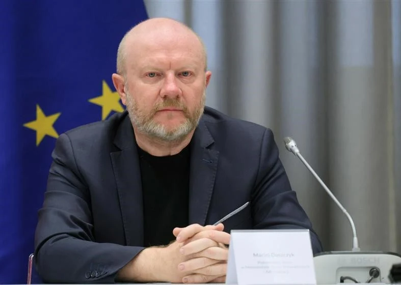 Mężczyzna z brodą ubrany w ciemny garnitur siedzi przy stole konferencyjnym, trzymając długopis, w tle widoczna flaga Unii Europejskiej oraz mikrofon.