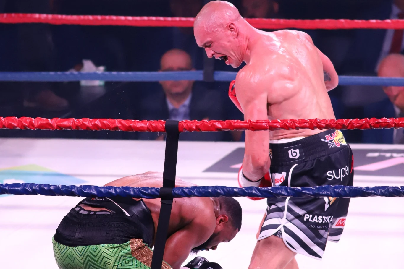 Dwóch bokserów w ringu podczas walki, jeden z zawodników klęczy, drugi stoi nad nim z uniesioną pięścią, otoczeni są linami ringu, w tle widoczna publiczność.