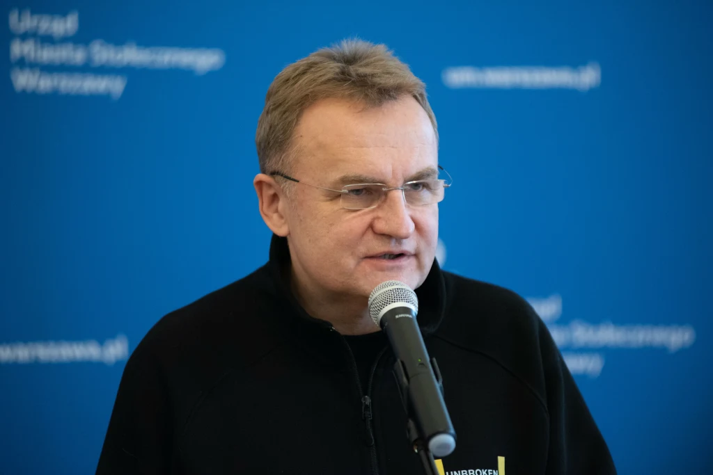 Mężczyzna w okularach mówi do mikrofonu, ubraną w ciemną kurtkę, na tle niebieskiej tablicy z napisem 'Urząd Miasta Stołecznego Warszawy'.