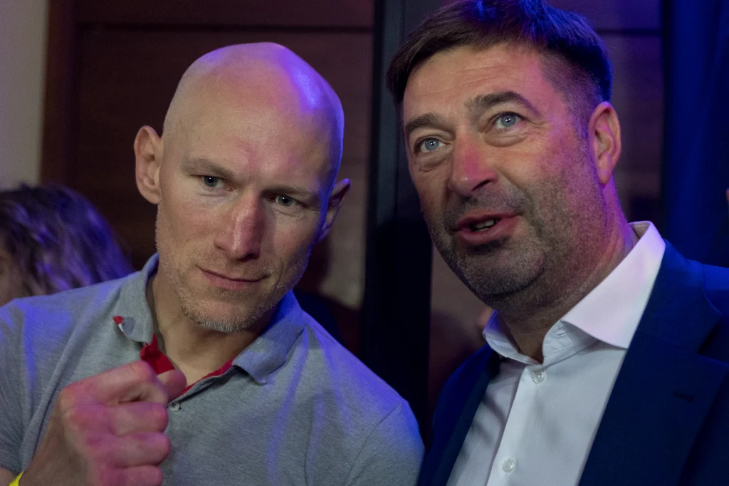 Krzysztof "Diablo" Włodarczyk i Andrzej Wasilewski Dwóch mężczyzn stoi blisko siebie w dobrze oświetlonym pomieszczeniu, jeden z nich wykonuje gest zaciśniętej pięści, obaj patrzą w tym samym kierunku, mają skupione wyrazy twarzy.