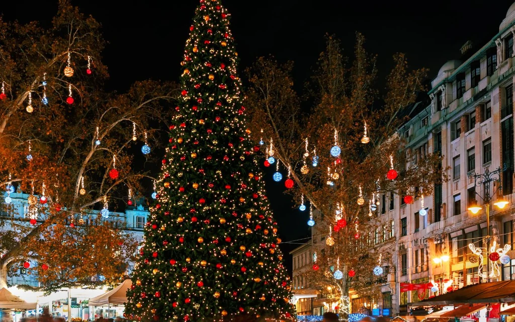 Udekorowana światełkami i bombkami choinka otoczona rozświetlonymi drzewami oraz kamienicami, tłum ludzi uczestniczy w świątecznym jarmarku nocą.