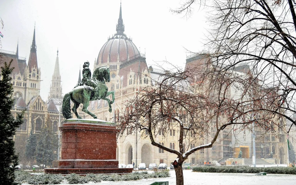 Zasypana śniegiem statua jeźdźca na koniu na tle imponującego gmachu parlamentu w Budapeszcie, otoczona drzewami i zimowym krajobrazem.