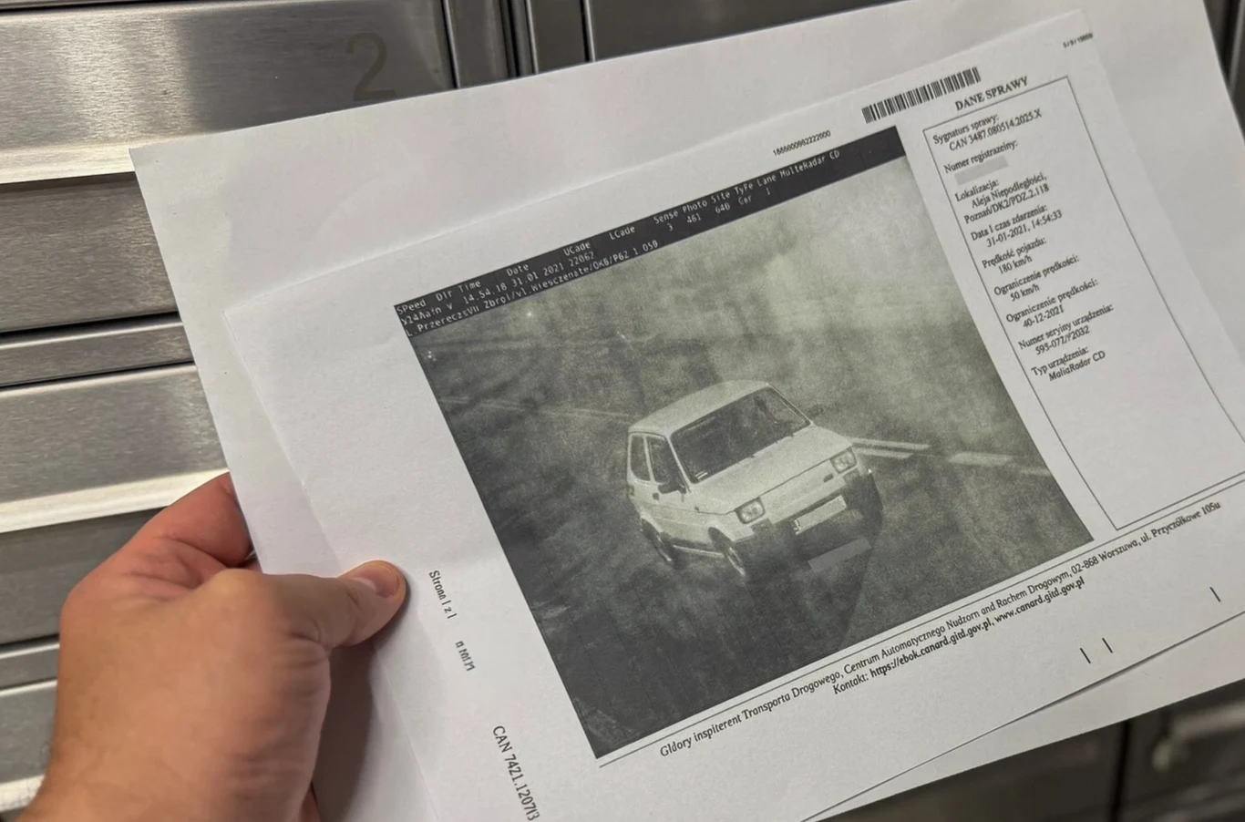 Nie odebrałem listu z fotoradaru – co z mandatem? Ręka trzyma dokument zawierający wydruk zdjęcia samochodu uchwyconego przez kamerę monitoringu, na tle metalowych szafek lub skrzynek pocztowych. Najważniejszym elementem jest czarno-białe zdjęcie auta oraz fragmenty danych widoczne na wydruku.