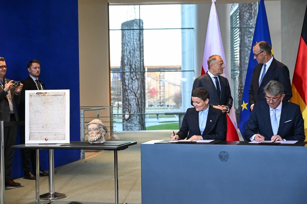 Niemcy. Zrabowane dokumenty wrócą do Polski. Ministerstwo kultury ogłasza Delegacje Polski i Niemiec podpisujące dokumenty przy stole, w tle widoczne flagi obu państw oraz Unii Europejskiej. Po lewej stronie na osobnym stoliku umieszczony został historyczny dokument i artefakt. W tle kilku mężczyzn w garniturach obserwuje wy...