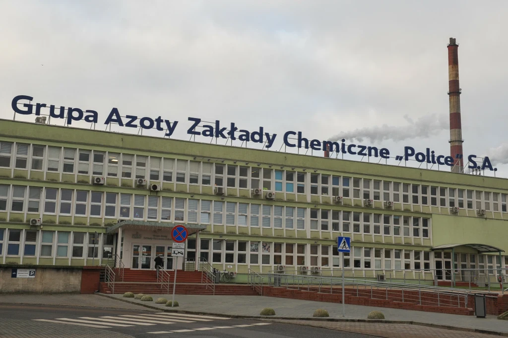 Fasada wielopiętrowego budynku przemysłowego z napisem Grupa Azoty Zakłady Chemiczne Police S.A., na pierwszym planie chodnik, schody oraz elementy infrastruktury miejskiej, w tle komin przemysłowy.