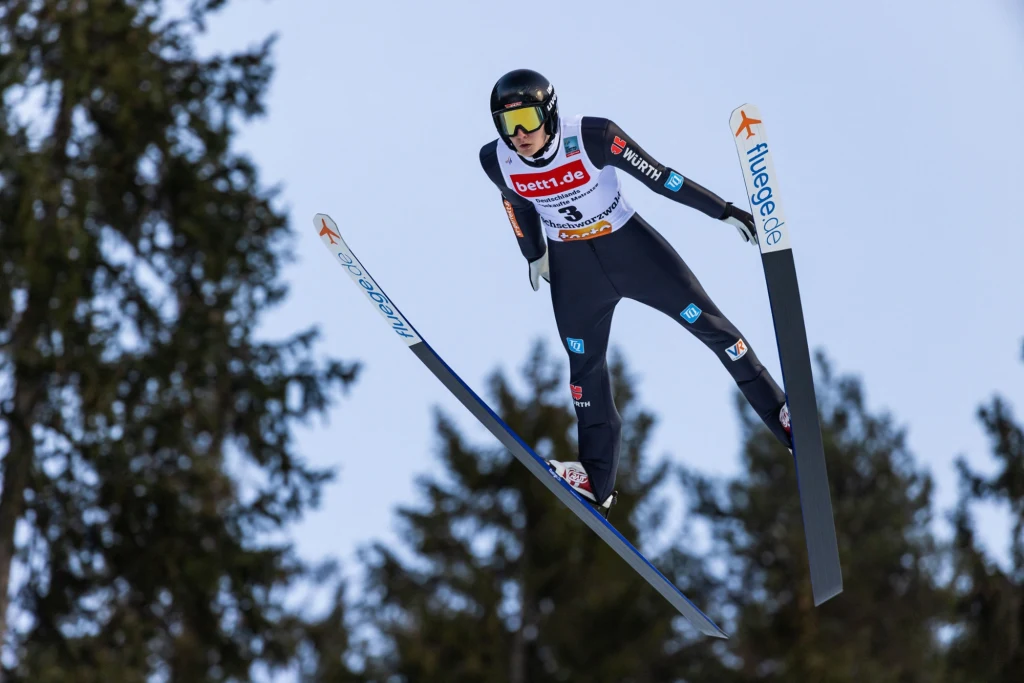 Justin Lisso Zawodnik w stroju narciarskim wykonuje skok podczas konkursu skoków narciarskich, znajdując się w powietrzu nad iglastymi drzewami, z widocznym numerem startowym i sponsorami na kombinezonie.