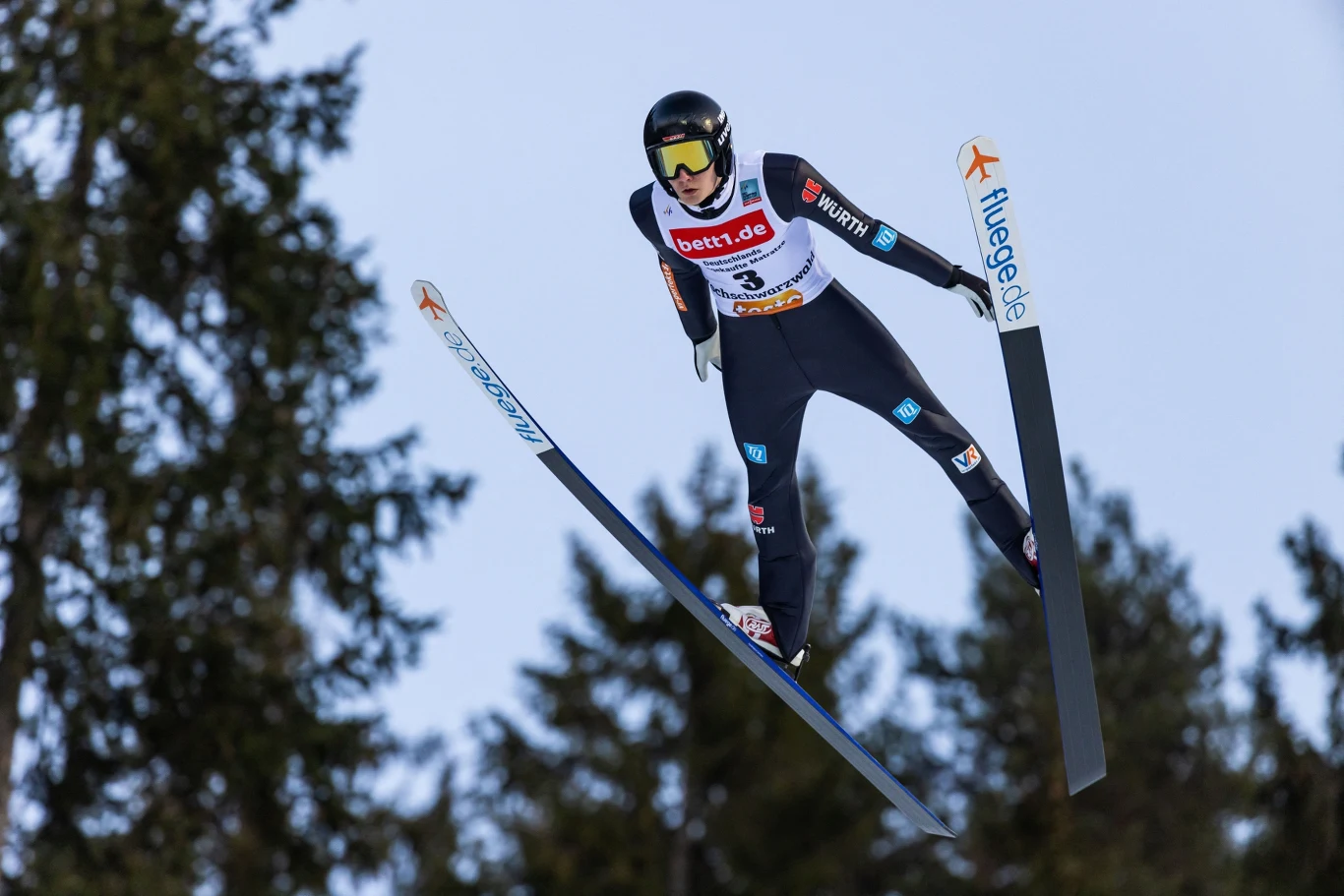 Zawodnik w stroju narciarskim wykonuje skok podczas konkursu skoków narciarskich, znajdując się w powietrzu nad iglastymi drzewami, z widocznym numerem startowym i sponsorami na kombinezonie.
