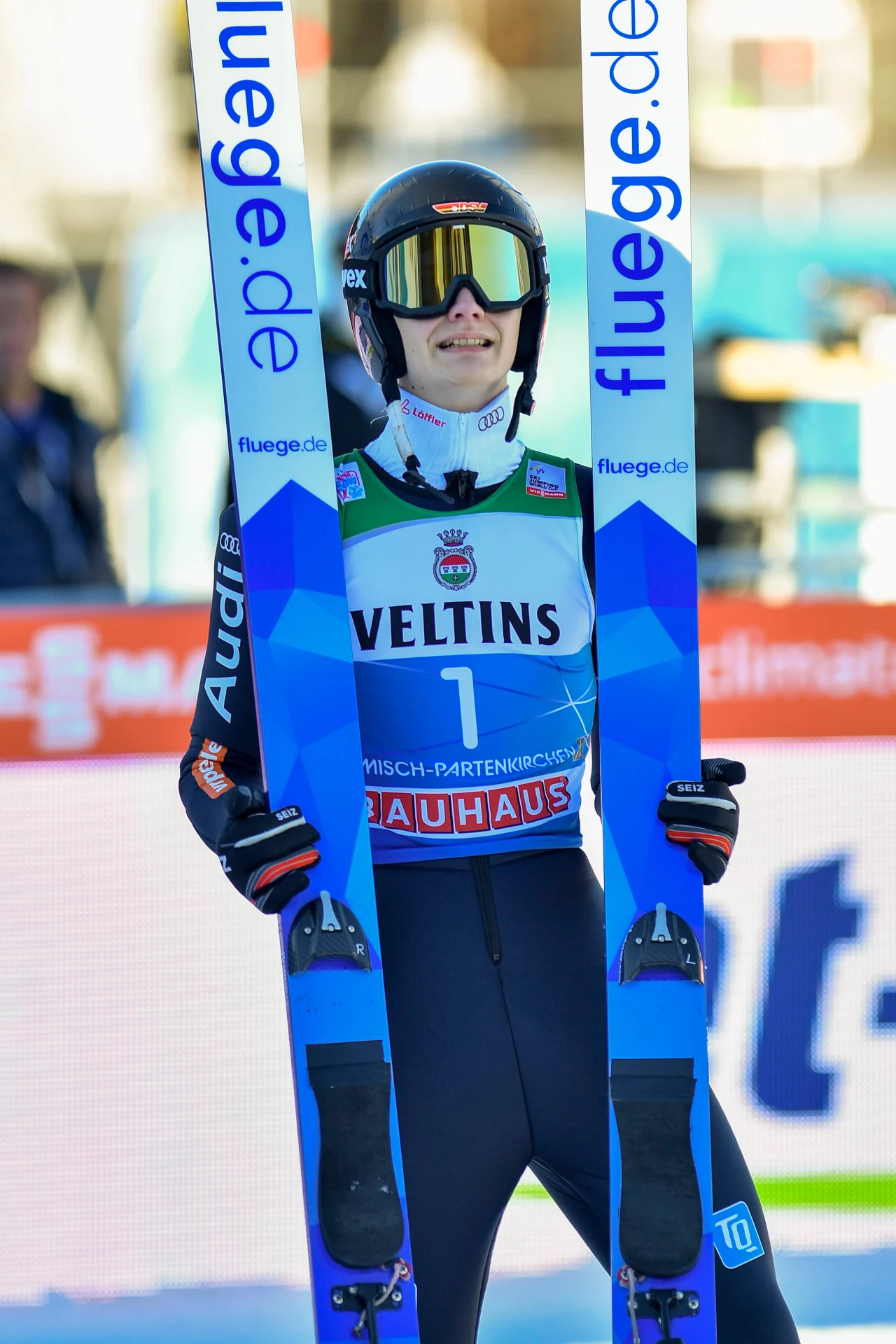 Justin Lisso Sportowiec w kombinezonie narciarskim i kasku trzyma narty do skoków narciarskich, stojąc na tle banerów z logo sponsorów. Uśmiechnięta twarz i numer jeden na koszulce startowej podkreślają sukces lub udział w zawodach.