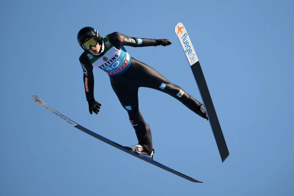 Justin Lisso Zawodnik w kombinezonie narciarskim i kasku wykonuje skok narciarski w powietrzu na tle błękitnego nieba, dynamiczna poza podkreśla moment lotu.
