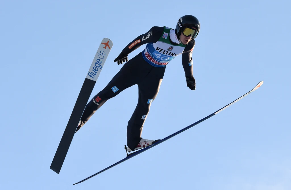 Justin Lisso Zawodnik w kombinezonie sportowym i kasku podczas skoku narciarskiego w powietrzu, dynamiczne ujęcie na tle błękitnego nieba