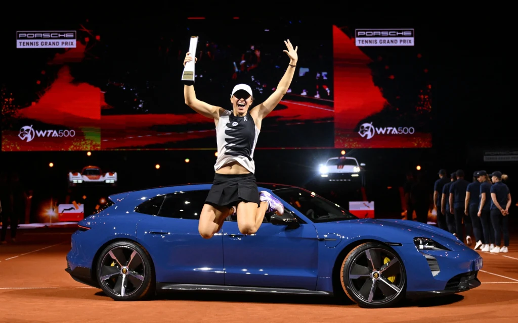 Zwyciężczyni turnieju tenisowego świętuje sukces, siedząc na niebieskim samochodzie sportowym z uniesionymi rękami i pucharem w dłoni. W tle ekrany z logotypami WTA500 i Porsche Tennis Grand Prix oraz grupa osób stojących na kortach.