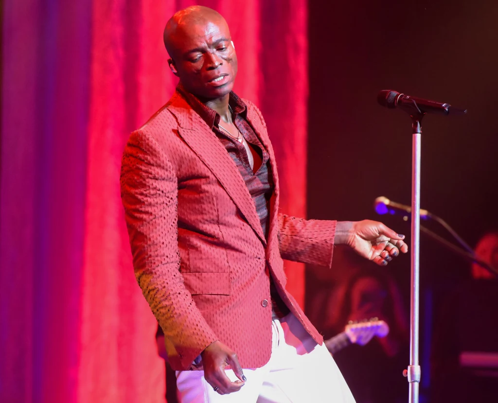 Mężczyzna ubrany w czerwony garnitur stoi na scenie przy mikrofonie, w tle czerwone zasłony i reflektory oświetlające przestrzeń.