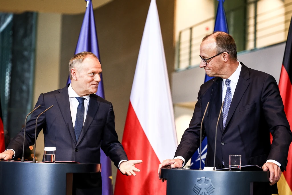 Dwóch mężczyzn w garniturach stoi za mównicami podczas konferencji prasowej. W tle widoczne są flagi Polski, Niemiec, Unii Europejskiej oraz NATO, podkreślające oficjalny charakter wydarzenia.