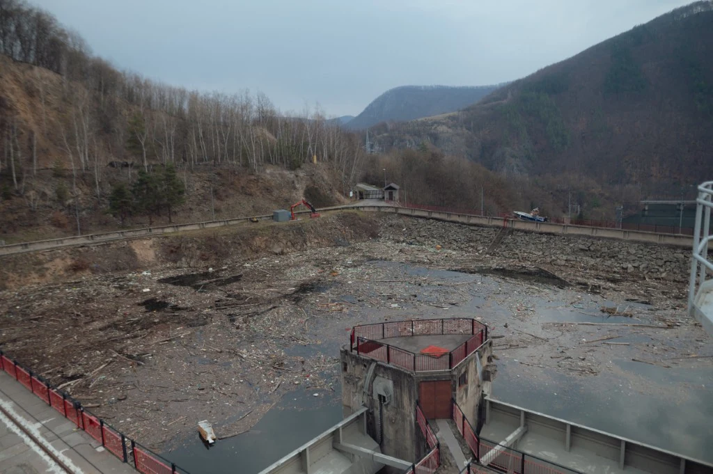 Tony odpadów zasypały słowackie jezioro Zaniedbany zbiornik wodny z widocznymi resztkami śmieci i gałęzi unoszącymi się na powierzchni, otoczony ogrodzeniem, w tle wzgórza oraz rzadko porośnięty las, szare niebo podkreśla ponury nastrój miejsca