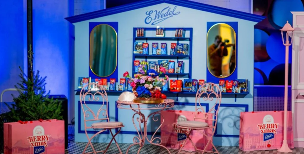 Niebieska aranżacja sklepu firmowego E. Wedel z ekspozycją słodyczy na półkach, dwiema owalnymi lustami, dekoracyjnym stołem z kwiatami oraz dwoma różowymi krzesłami, otoczona różowymi pudełkami promocyjnymi Berry Times.